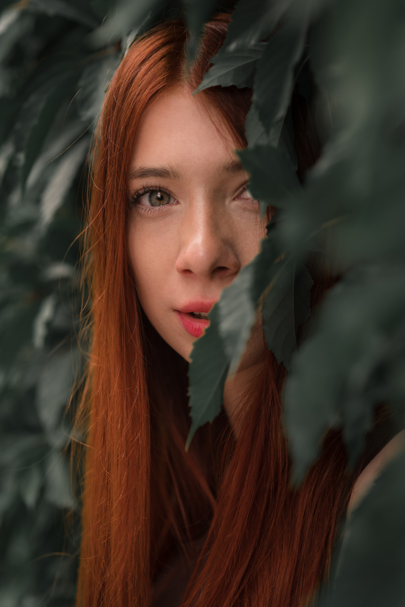 Klimatyczny portret rudowłosej dziewczyny w otoczeniu zieleni, kadr z bliska z naturalnym ramowaniem przez liście